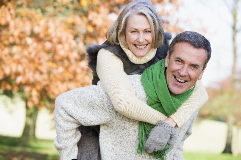 A older couple out in a park. The woman is hanging on to the guy's back, like a backpack.